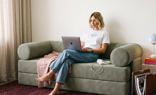 Woman sitting cross-legged on a textured green sofa, working on a laptop in a bright, cozy living room.