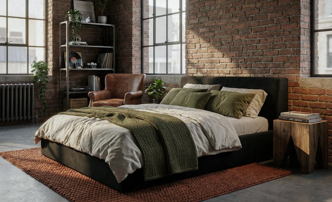 # Front view of O2 Soft Frame Bed in black, featuring minimalist bedding, a cozy throw, and a wooden side table. The industrial-style room is complemented by natural light, exposed brick walls, and greenery.