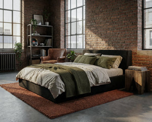 # Front view of O2 Soft Frame Bed in black, featuring minimalist bedding, a cozy throw, and a wooden side table. The industrial-style room is complemented by natural light, exposed brick walls, and greenery.