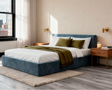 O2 Bed Cloud Frame in deep blue, styled with white bedding, olive green pillows and a cozy throw, set in a warm modern bedroom with wood nightstands and large windows.