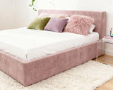 O2 Bed Cloud Frame in soft pink, styled with white bedding, pastel pillows, and a fluffy throw, set in a bright, cozy bedroom with a plush rug and modern acrylic nightstand.