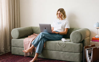 Woman sitting cross-legged on a textured green sofa, working on a laptop in a bright, cozy living room.