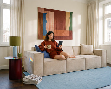 Woman relaxing on a beige modular sofa with a mug and tablet in a sunlit living room.