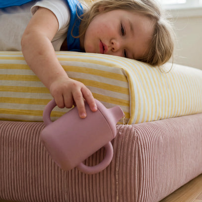 A young child lies on a bed with yellow-and-white striped bedding, reaching toward a dusty pink silicone teapot-shaped cup resting on the bed.