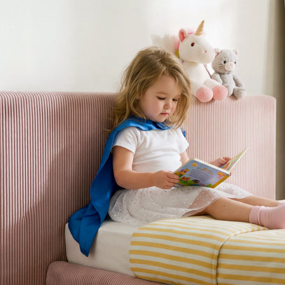 A young girl with blonde hair sits on a pink ribbed sofa, reading a colorful book. She is wearing a white t-shirt, a white polka-dot skirt, and a blue cape.