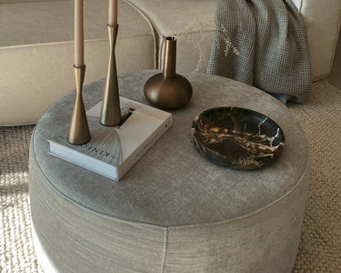 #A gray round pouf ottoman  in a cozy living room with minimal decor, featuring two candle holders, a vase, and a marble tray