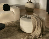 #A soft-textured ottoman in a neutral living room with a cozy blanket and a glass of water on top, next to a beige modular sofa